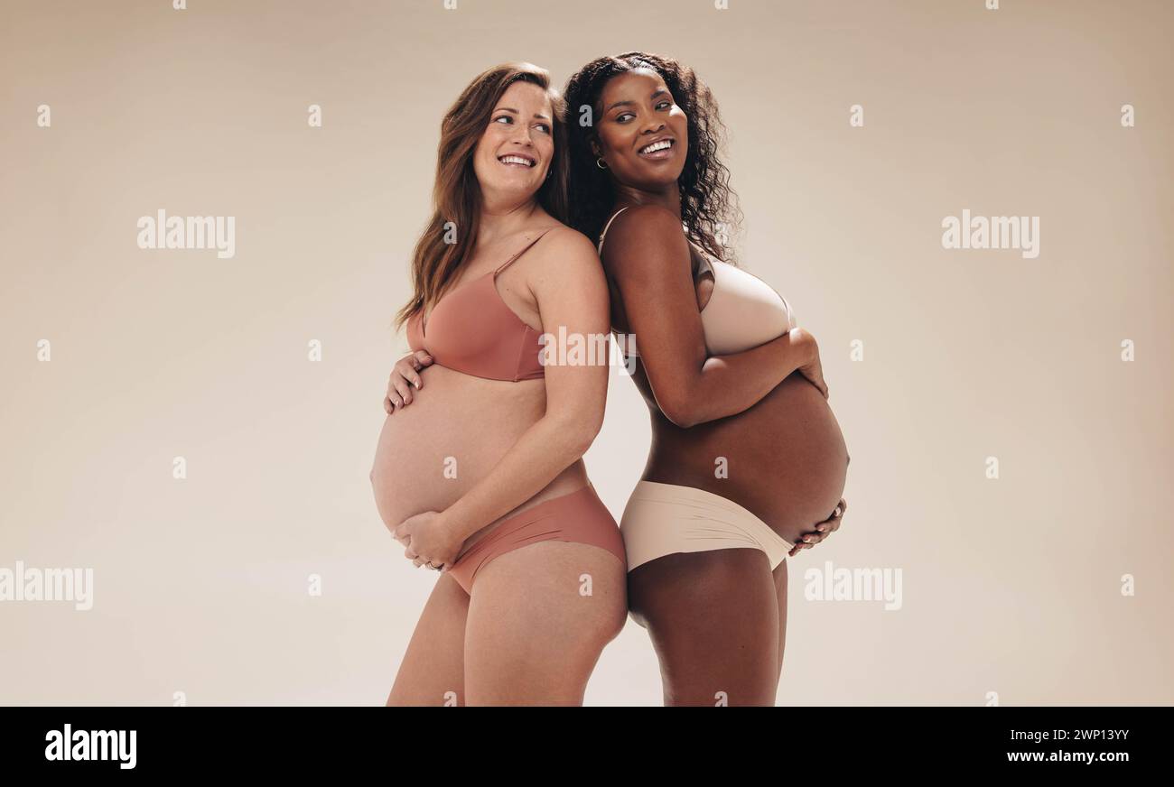 Zwei selbstbewusste, schwangere Frauen stehen zusammen in einem Studio. Sie zeigen stolz ihre Babybeulen, lächelnd und strahlend glücklich. Diese werdenden Mütter Stockfoto