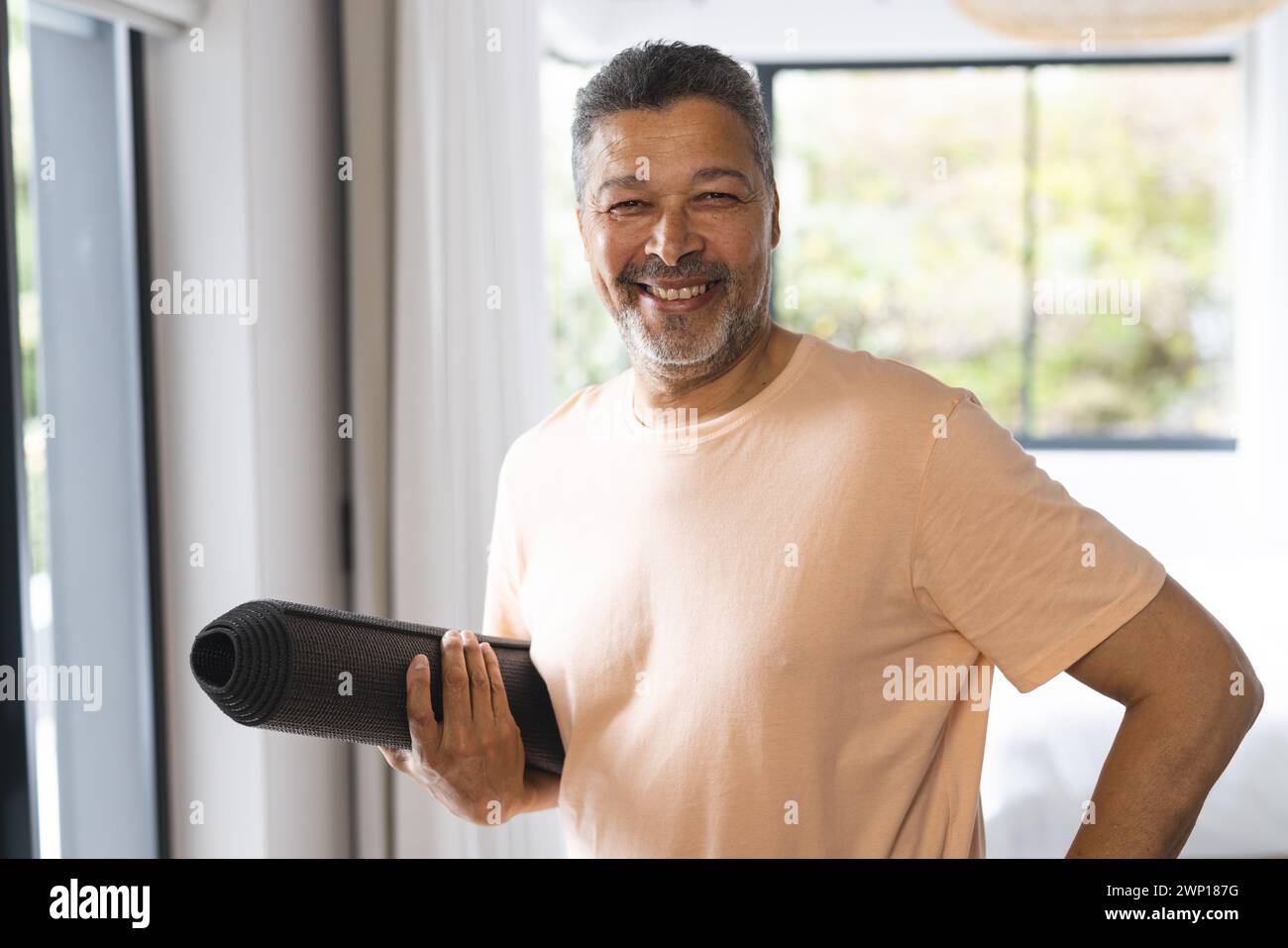 Ein birassischer Seniorenmann mit einer Yogamatte lächelt selbstbewusst und trägt ein lässiges pfirsichfarbenes Hemd Stockfoto