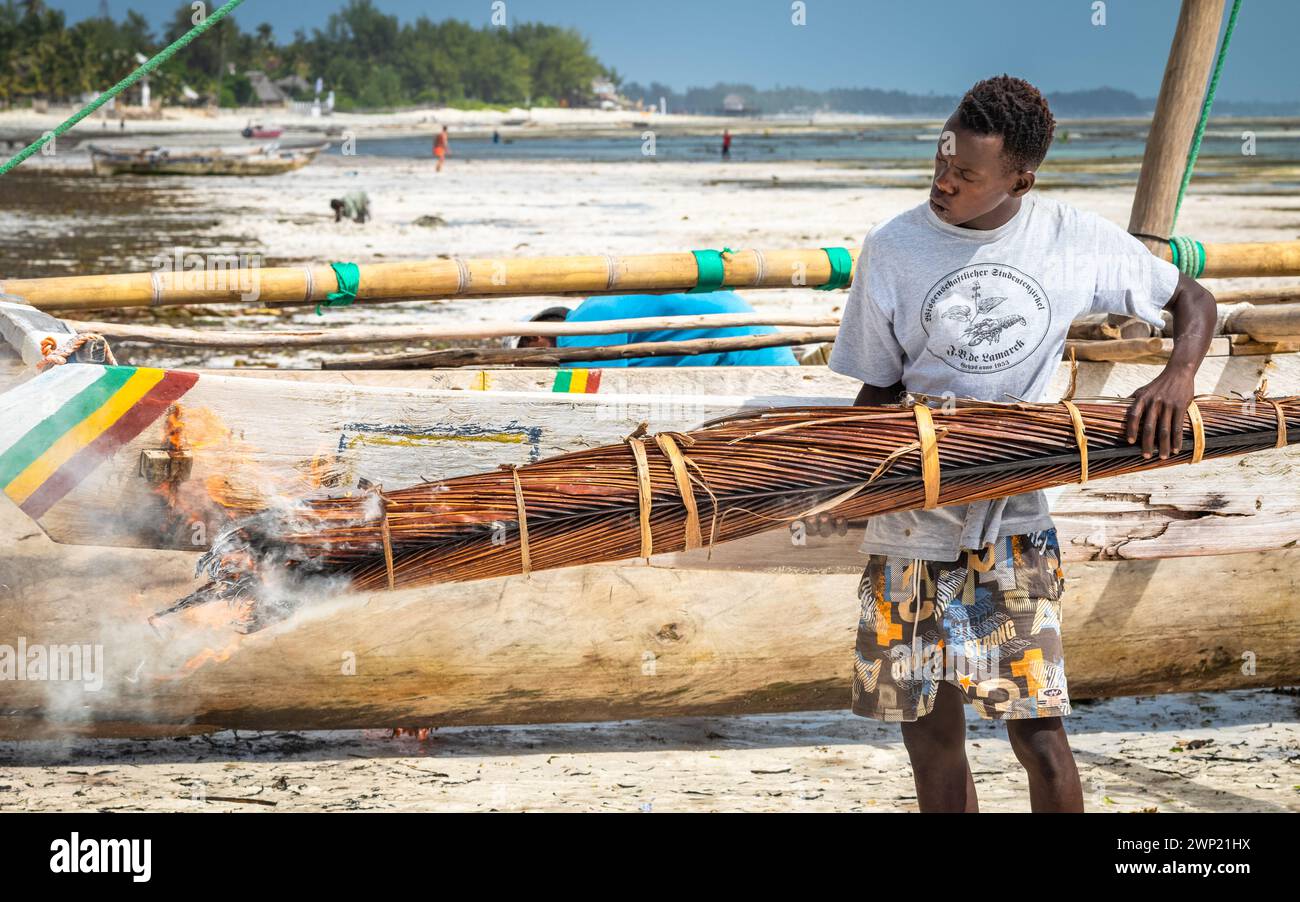 Ein Mann benutzt brennende Palmwedel, um sein traditionelles hölzernes Dhow-Boot, Jambiani, Sansibar, Tansania, zu verkohlen und zu wasserdicht zu machen Stockfoto