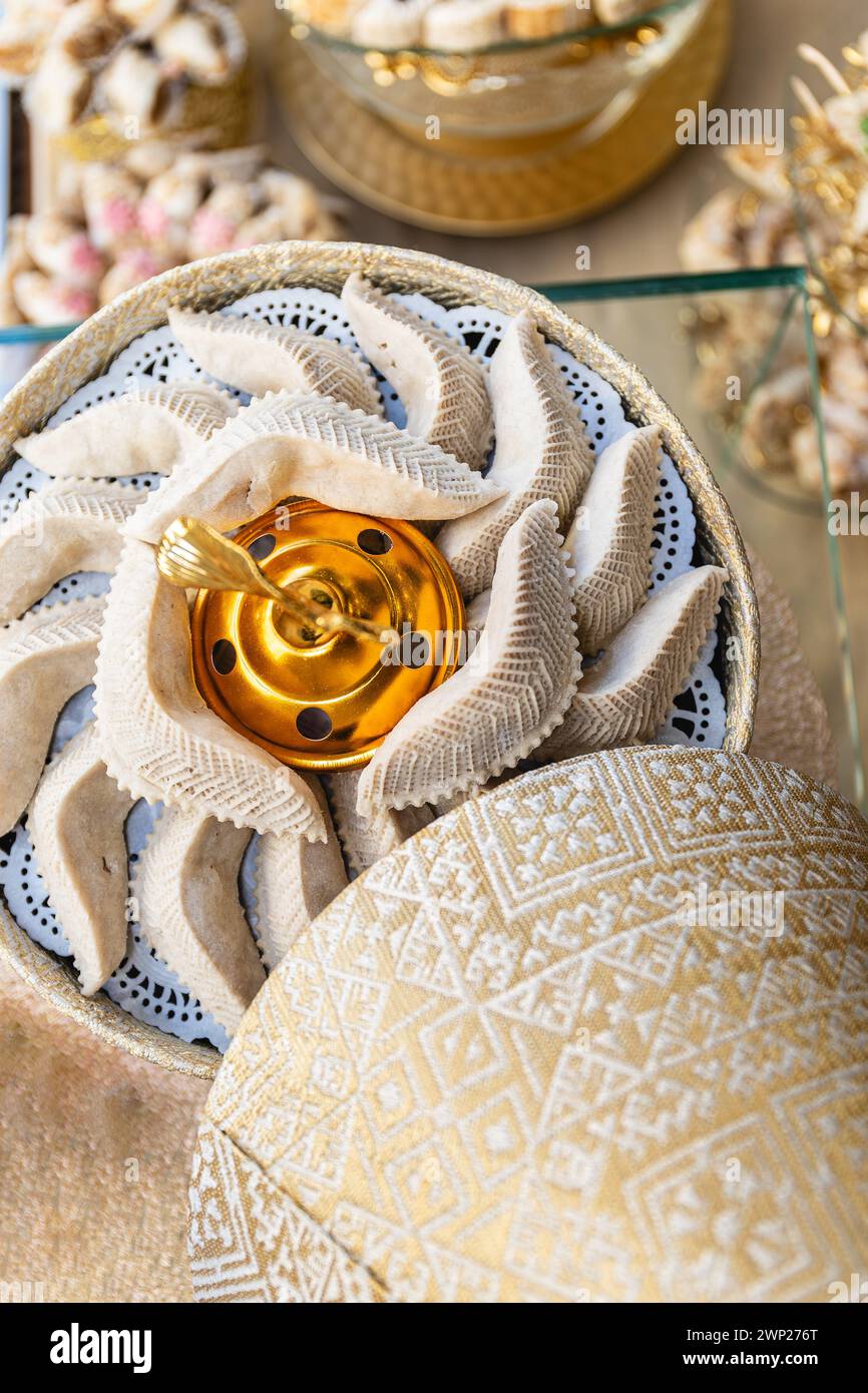 Vertikales Foto ein kunstvolles goldenes Herzstück, umgeben von halbmondförmigen arabischen Süßigkeiten, präsentiert auf einem Tisch mit zarten Spitzendetails. Essen und Stockfoto