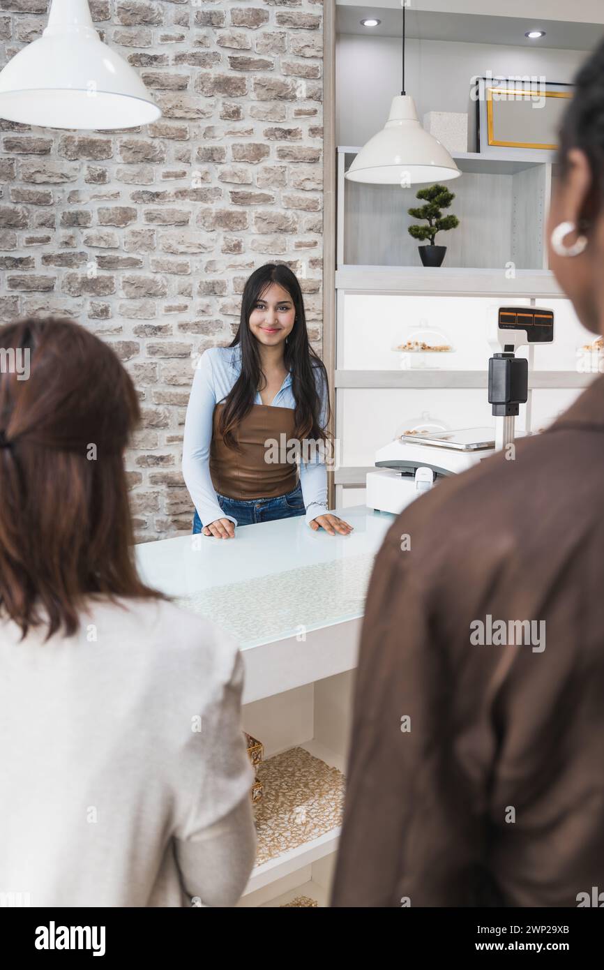 Ein vertikaler Foto lächelnder junger Bäckereibesitzer steht hinter der Theke bereit und begrüßt Kunden in einem modernen, stilvollen Bäckereigeschäft mit rustikaler Backsteinwand Stockfoto