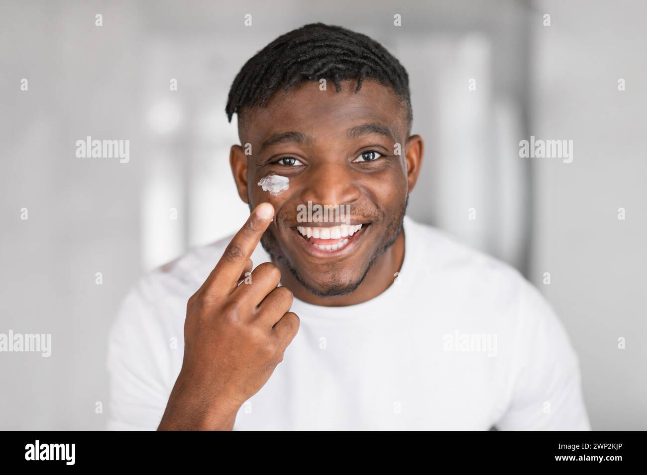 Der Kopfschuss des fröhlichen afrikanischen Mannes trägt Gesichtscreme auf die Wange auf Stockfoto