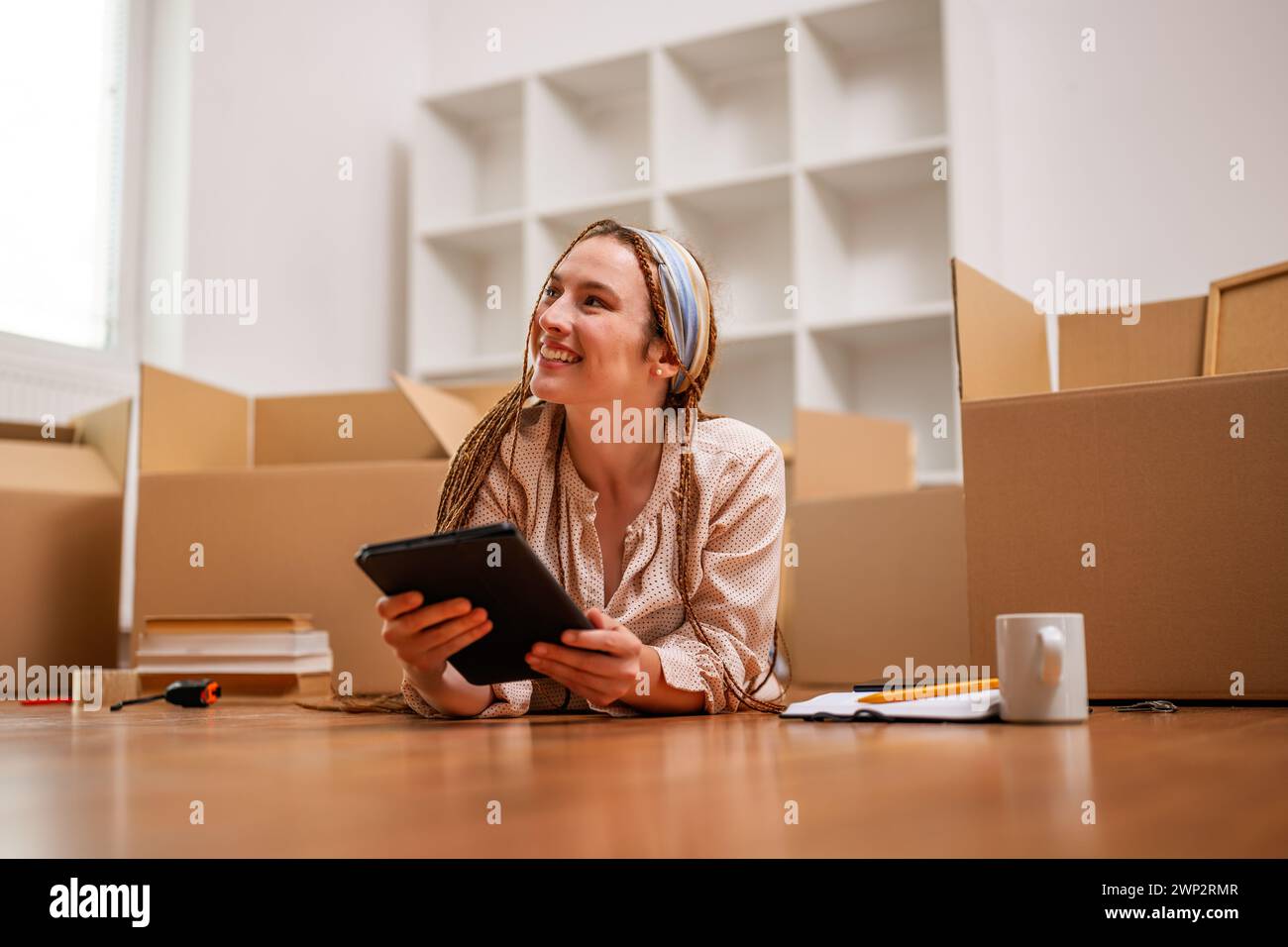 Moderne Ingwerfrau mit Zöpfen auf digitalem Tablet beim Einzug in eine neue Wohnung. Stockfoto