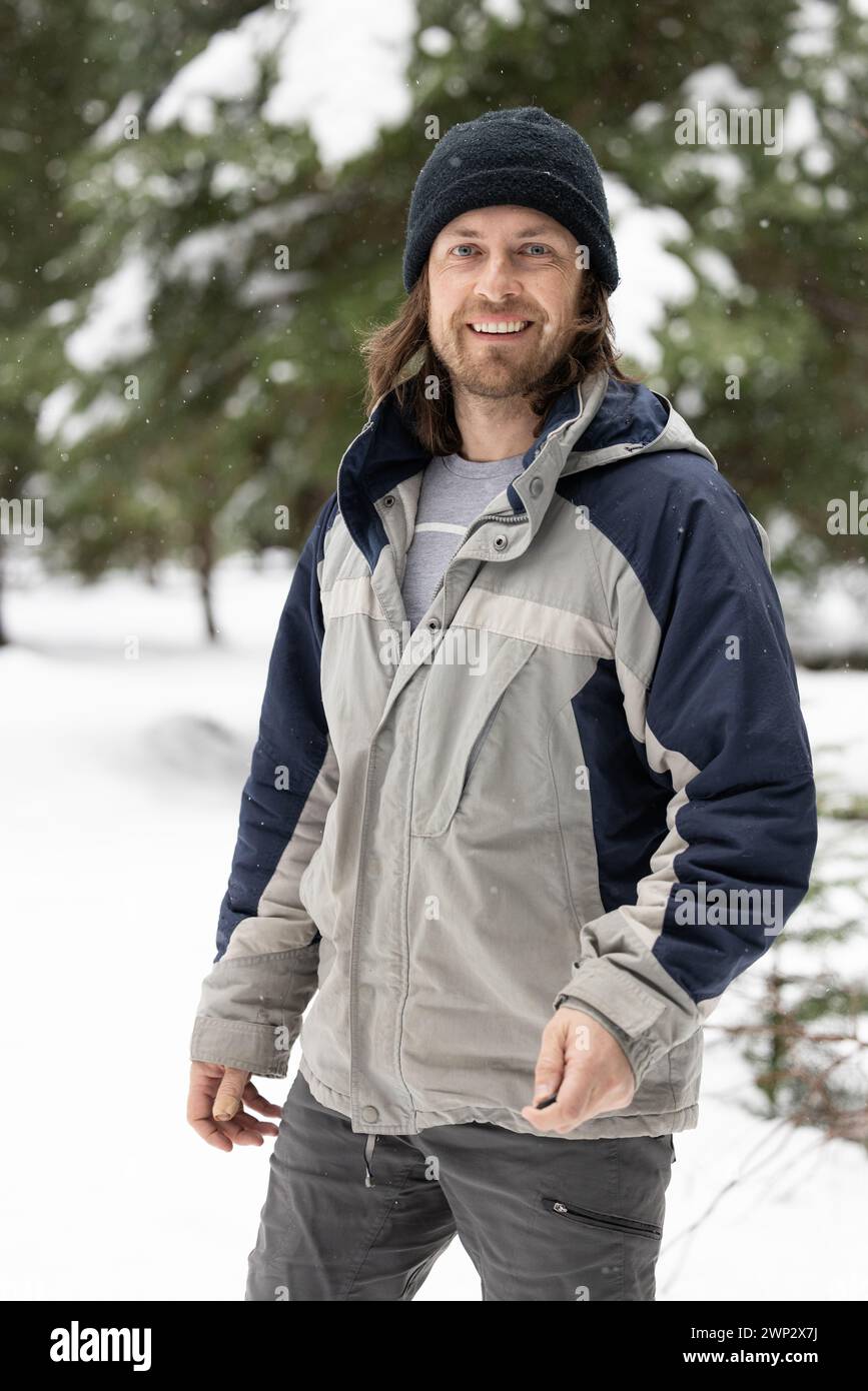 Ein Mann, der eine blau-graue Jacke und einen schwarzen Hut trägt, lächelt. Er steht im Schnee mit Bäumen im Hintergrund Stockfoto