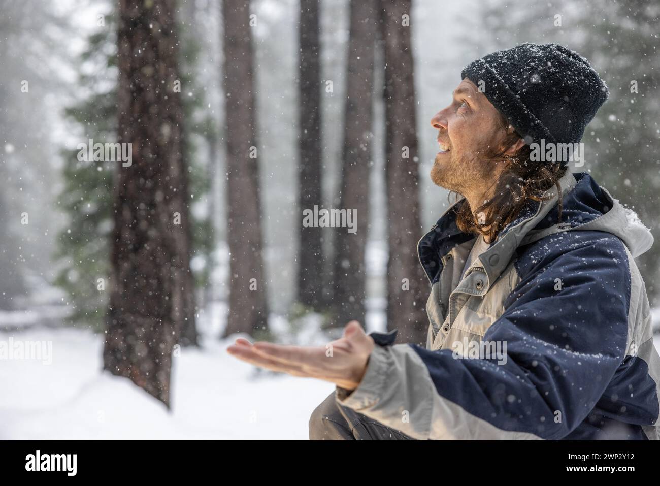 Ein Mann mit schwarzem Hut und blauer Jacke steht im Schnee und hält die Hände nach außen, um Schneeflocken zu fangen. Das Konzept von Freude und Verspieltheit als das Stockfoto