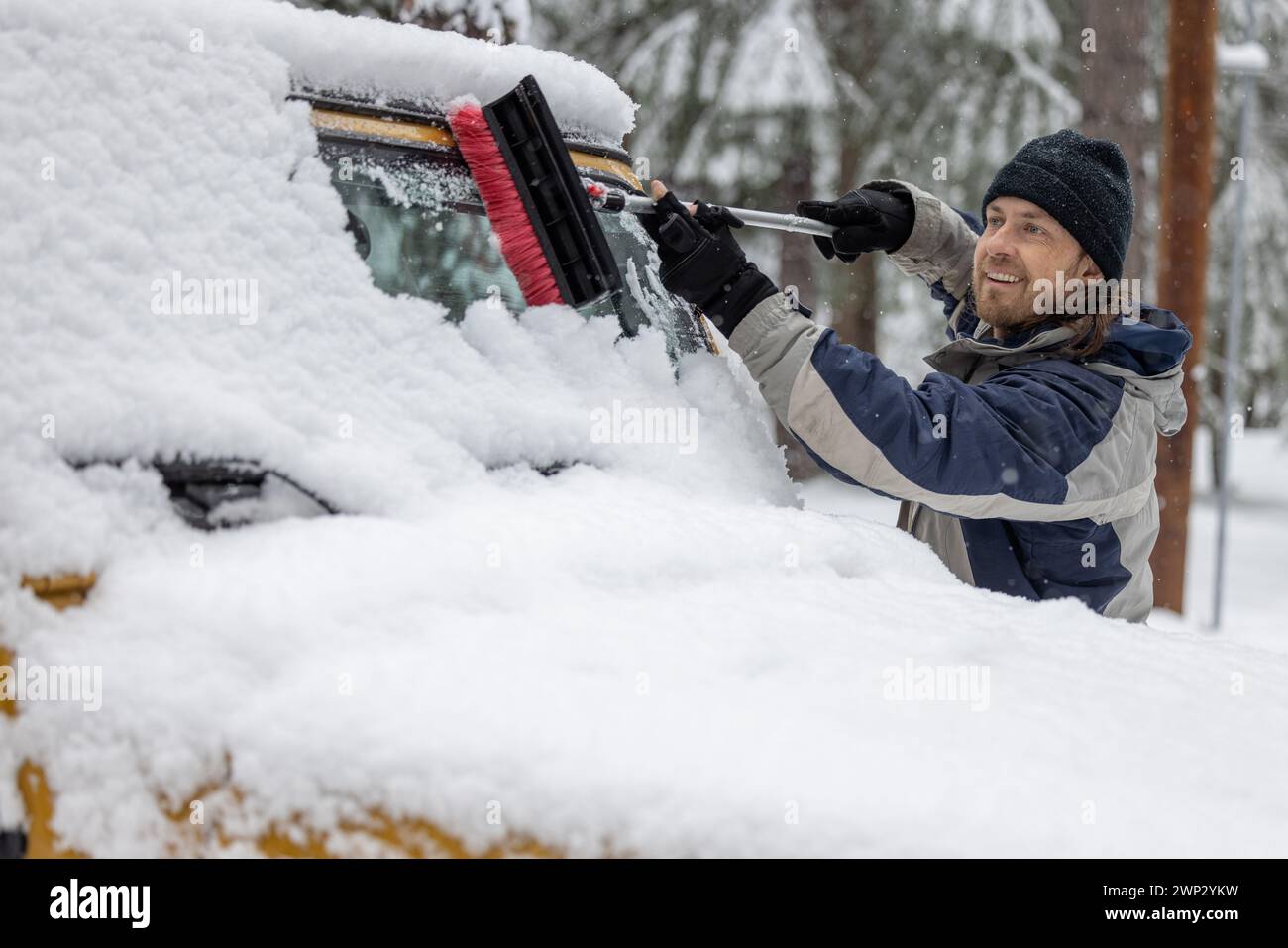 Ein Mann reinigt die Windschutzscheibe eines Autos mit einer Schneebürste. Der Mann lächelt und genießt die Aufgabe Stockfoto