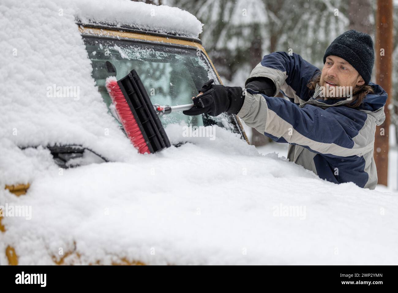Ein Mann räumt Schnee von einem Autofenster. Die Szene ist kalt und schneebedeckt, und der Mann trägt einen schwarzen Hut Stockfoto