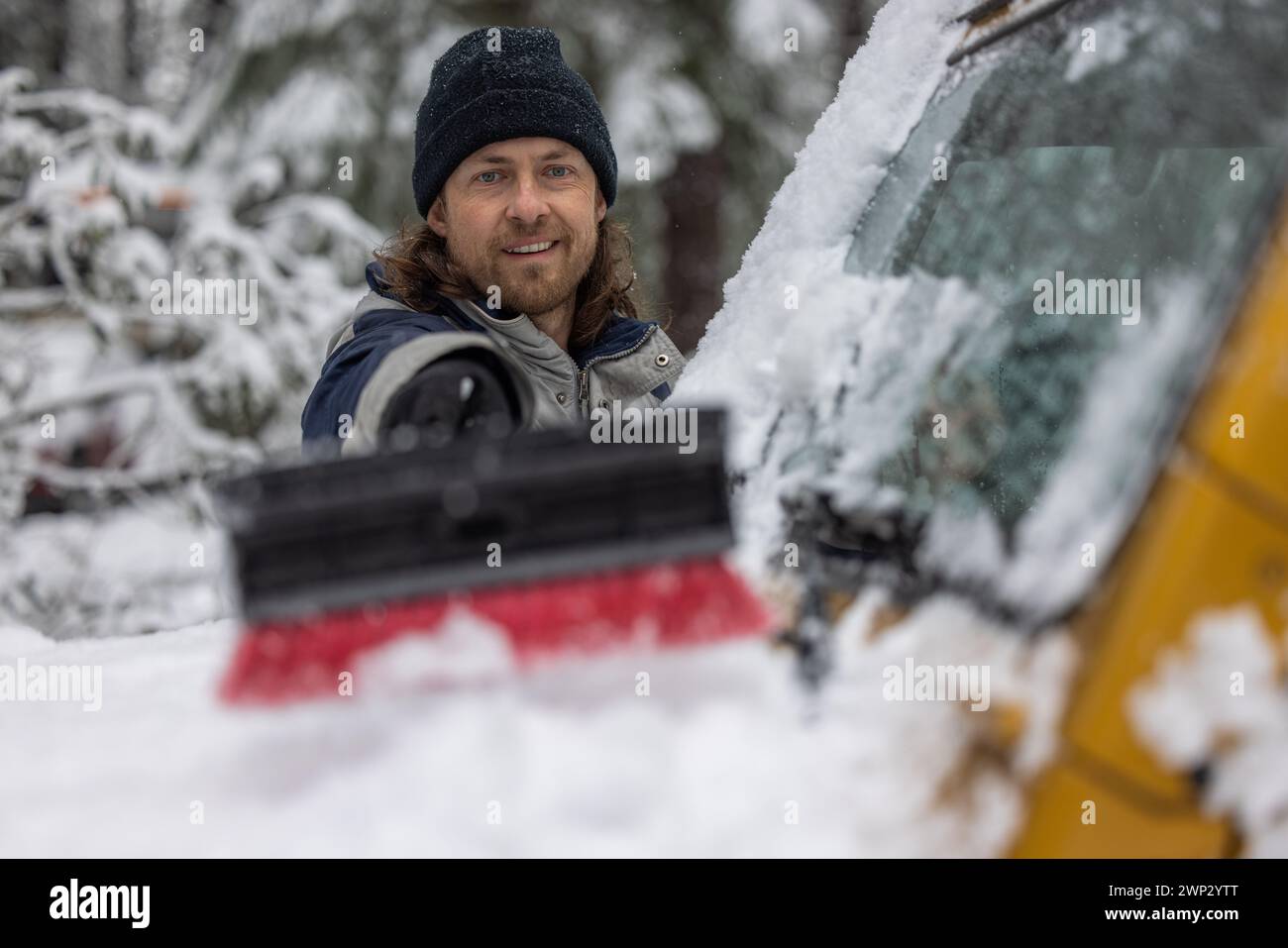 Ein Mann lächelt, während er Schnee von einem Auto schiebt. Die Szene spielt in einer verschneiten Umgebung, und der Mann trägt einen schwarzen Hut Stockfoto