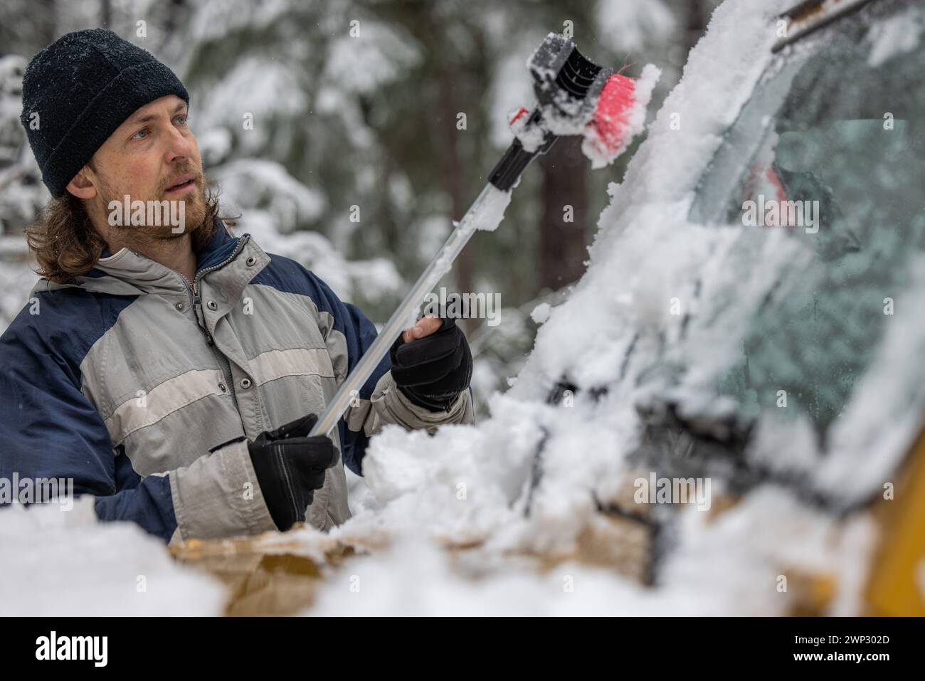 Ein Mann mit schwarzem Hut benutzt eine Schneebürste, um den Schnee von einem Auto zu entfernen Stockfoto