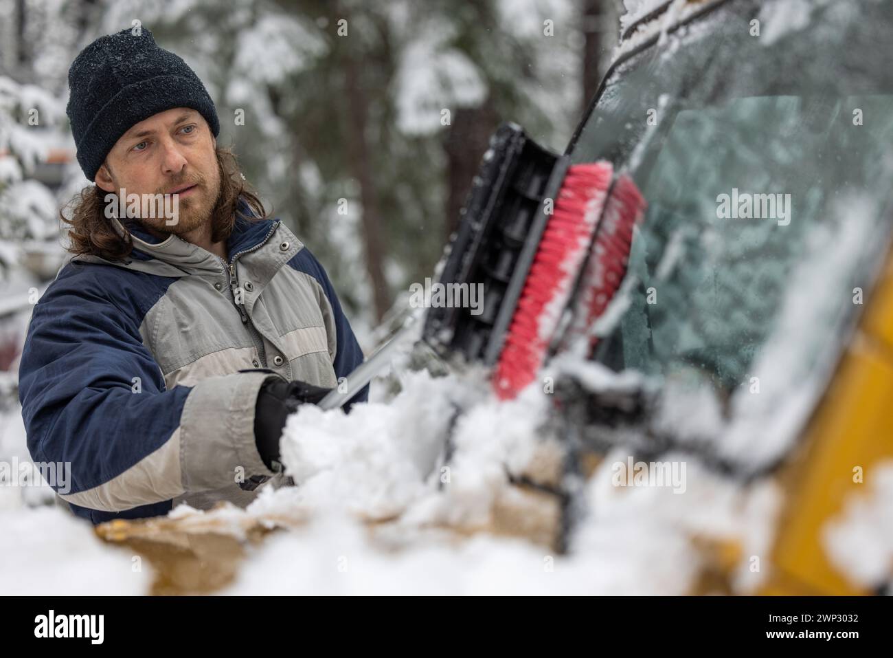 Ein Mann räumt Schnee von einem Autofenster. Der Mann trägt eine blaue Jacke und einen schwarzen Hut. Die Szene spielt in einer schneebedeckten Umgebung Stockfoto