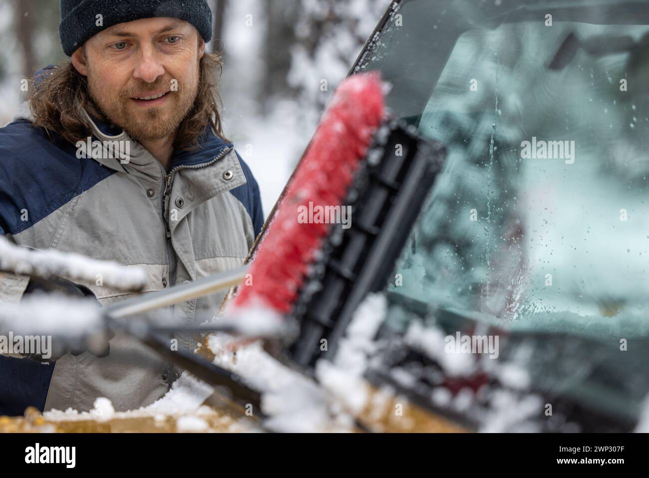 Ein Mann putzt sein Auto mit einer Schneebürste. Der Mann lächelt und genießt die Aufgabe Stockfoto