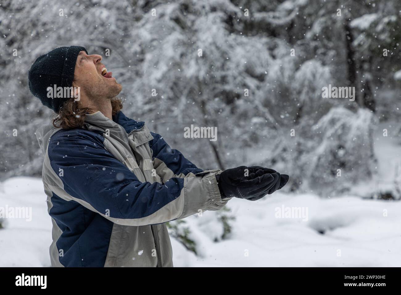 Ein Mann in einer blau-grauen Jacke wirft Schnee in die Luft. Er trägt einen schwarzen Hut und Handschuhe. Vorstellung von Freude und Verspieltheit, wie der Mann es genießt Stockfoto