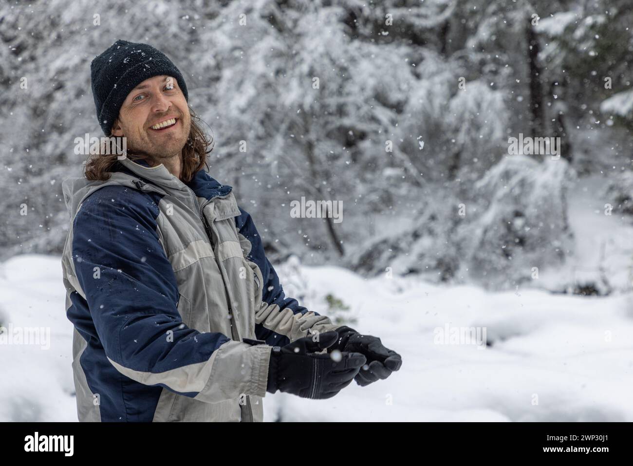 Ein Mann mit einer blau-grauen Jacke lächelt im Schnee. Er hält ein Paar Handschuhe und genießt das Winterwetter Stockfoto