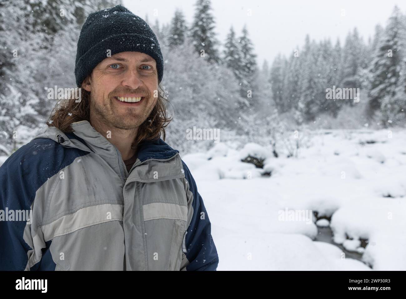 Ein Mann, der eine blau-graue Jacke und einen schwarzen Hut trägt, lächelt im Schnee Stockfoto