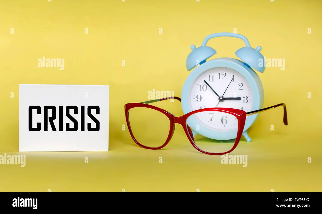 Notizblock mit Krisenzeichen, Geschäftsverlust, Krisenkonzept auf gelbem Hintergrund mit Wecker und Brille Stockfoto
