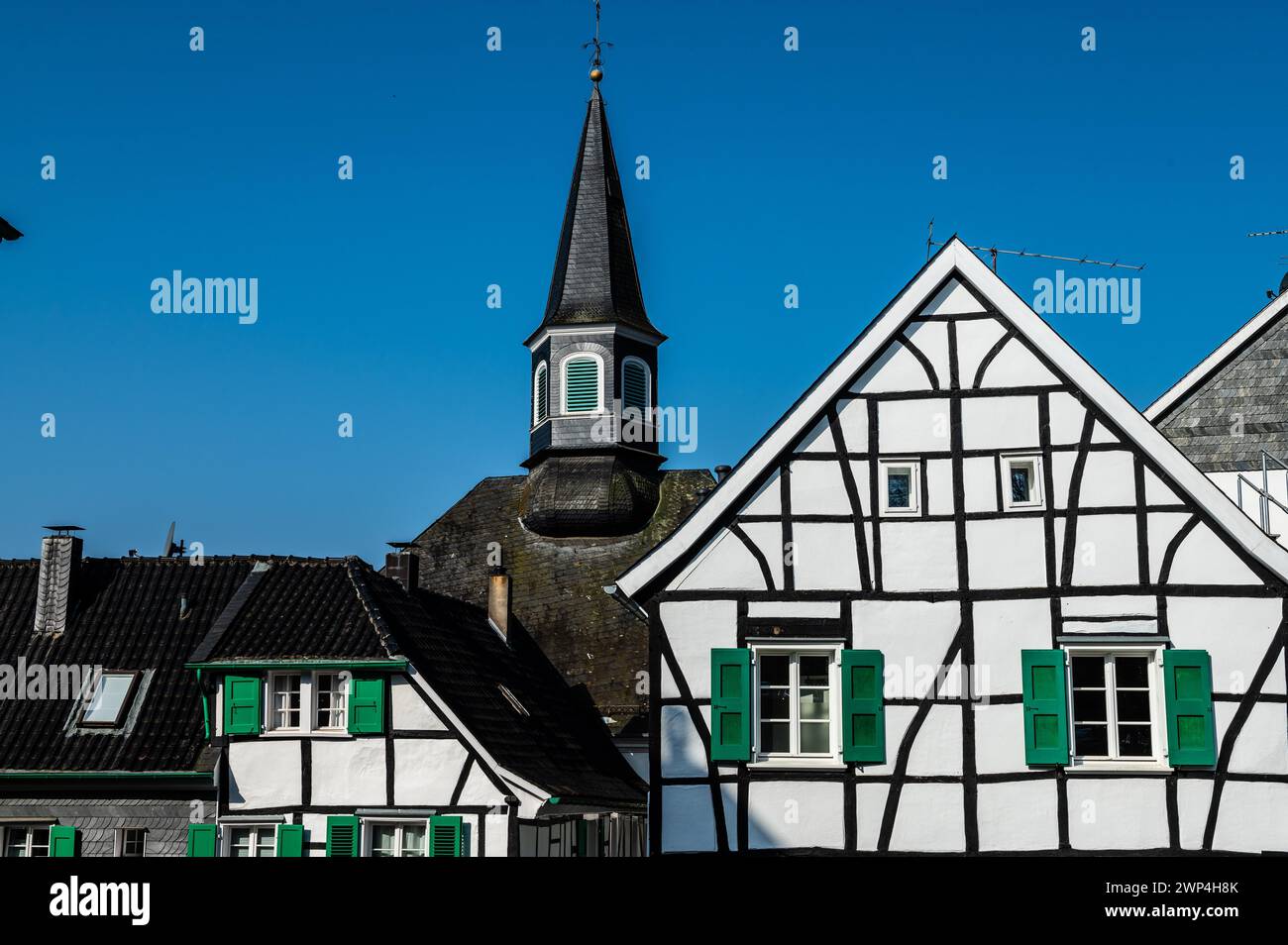 Weißes Fachwerkhaus mit grünen Fensterläden vor einem Kirchturm unter blauem Himmel, Graefrath, Solingen, Bergisches Land, Norden Stockfoto