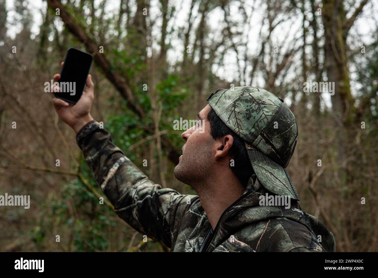 Ein Mann in Tarnung kämpft, um ein Handy zu finden, umgeben von dichtem Wald, mit einem Handy in der Hand Stockfoto