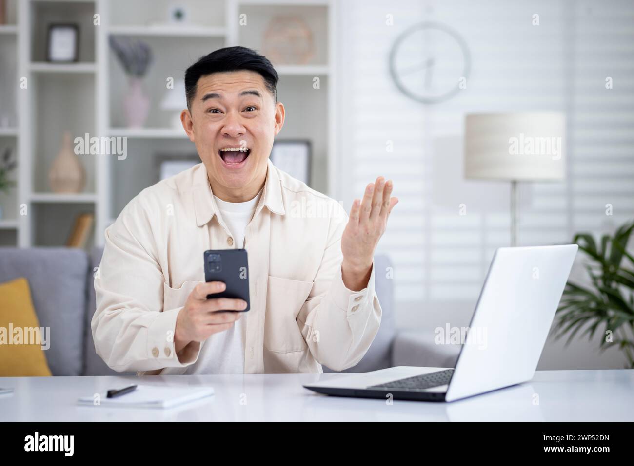 Staunte östliche Mann, der ein Mobiltelefon benutzte, während er an einem entfernten Arbeitsplatz mit PC und Notebook saß. Ekstatische Banktransaktionszahlung für Erwachsene, männliche Banktransaktionen auf digitalem Gerät während des Arbeitstages. Stockfoto