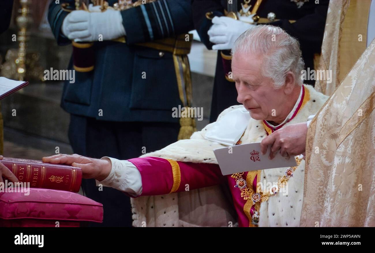 König Karl III., der in zeremoniellen Gewändern sitzt, legt während der Krönungszeremonie in Westminster Abbey Westminster London, UK, am 6. Mai 2023 den feierlichen Krönungseid ab, der die Heilige Bibel berührt, die mit seinem Herrscher Cypher und Datum eingraviert ist Stockfoto