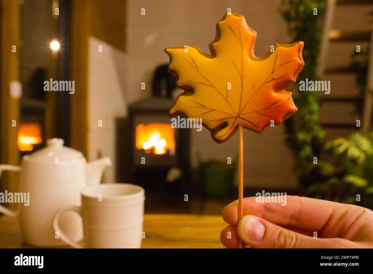 Ahornblatt-förmiges Ahornblatt in der Hand der Frau gegen brennendes Feuer im Kaminhintergrund. Am Herbstabend in gemütlichem Haus ausruhen. Weiße Teekanne und Kuh Stockfoto