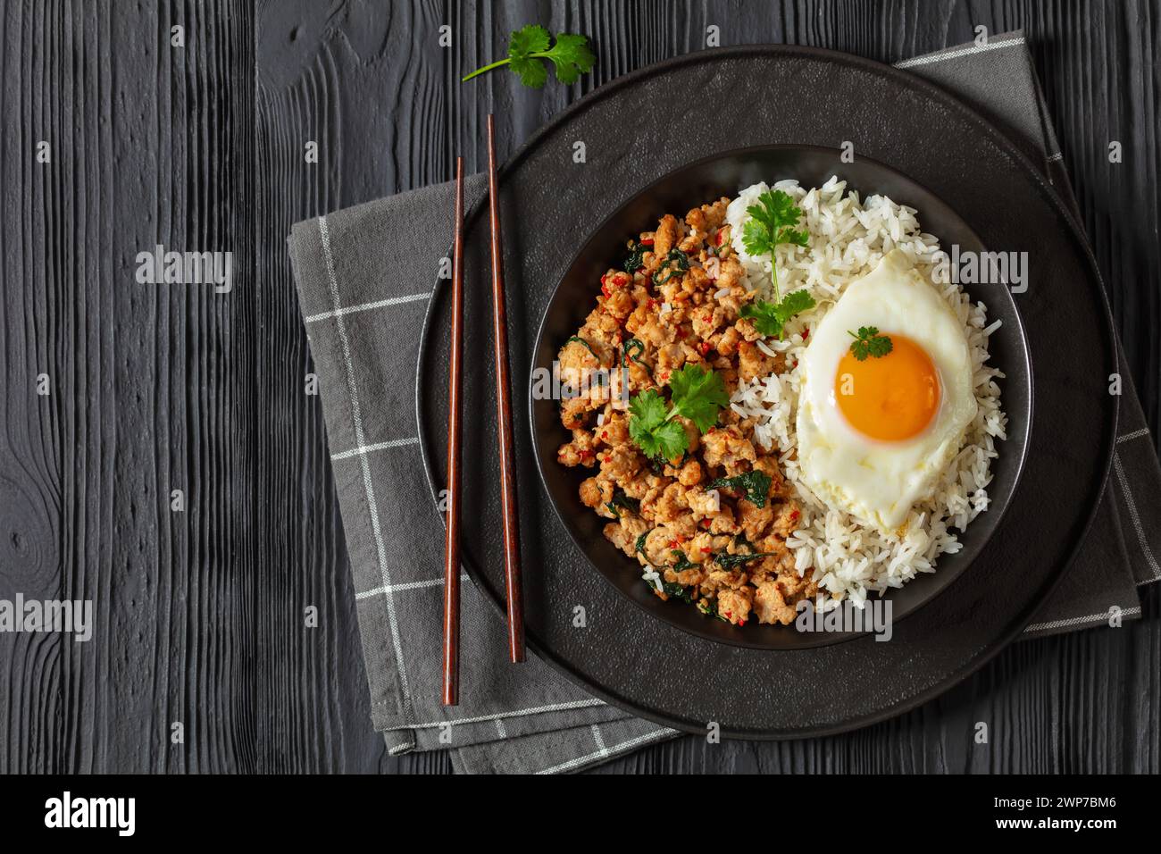 Pad Krapow Gai, Pad Gaprao, Pad kra Pao, Basilikum-Hühnchen-Rührbraten mit Reis und Spiegelei auf Teller mit Essstäbchen auf schwarzem Holztisch, thailändische Küche, Stockfoto