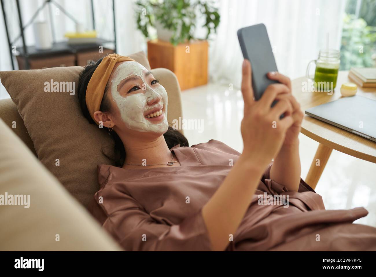 Glückliche Frau mit Tonmaske im Gesicht, die sich auf der Couch entspannt und Freunden SMS schickt Stockfoto