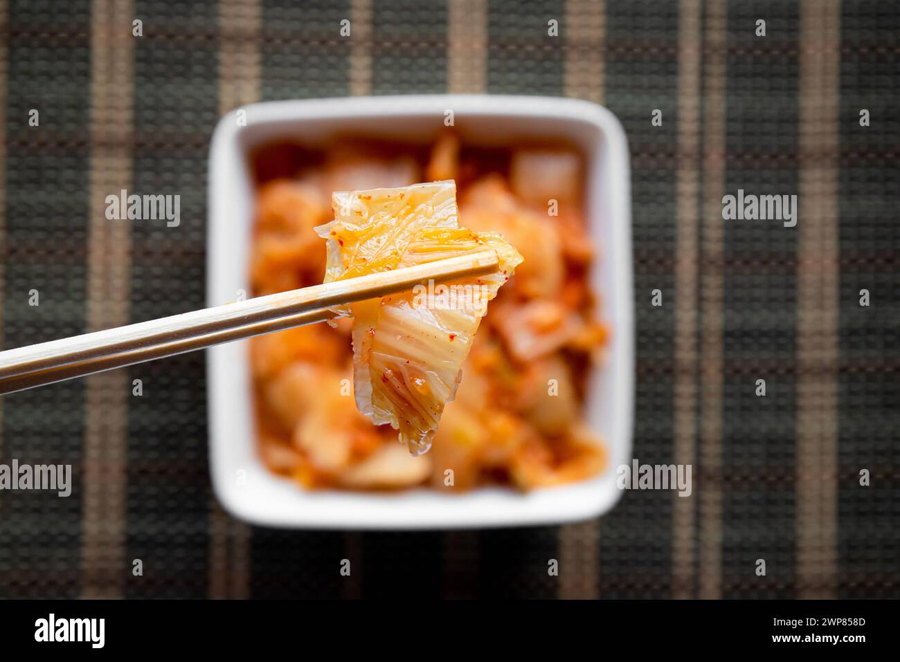 Selektiver Fokus auf Bambus-Essstäbchen mit fermentiertem Kimchi-Kohl-Salat und weißer Schüssel auf braunem Hintergrund. Stockfoto