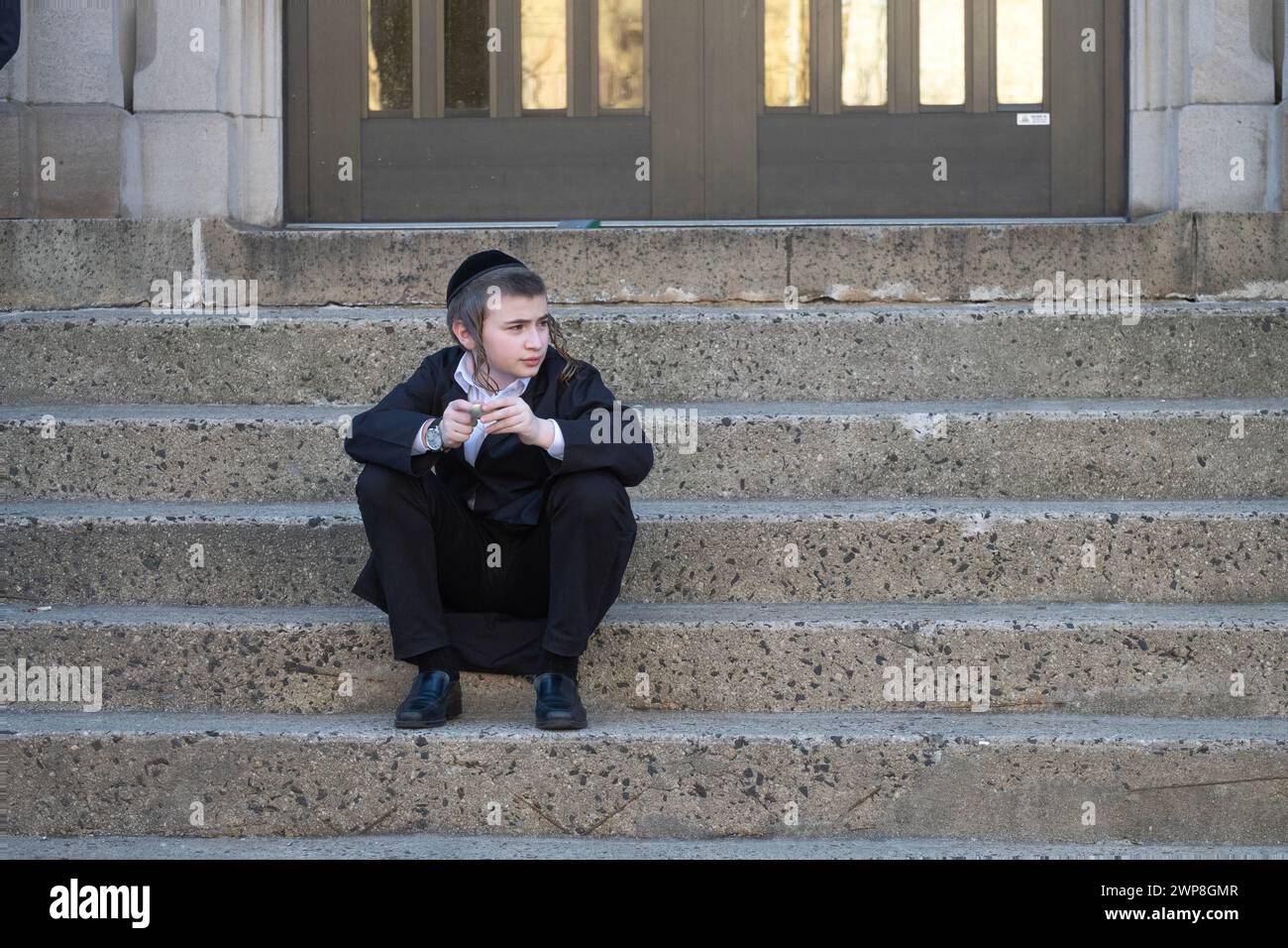 In der Pause sitzt ein junger orthodoxer jüdischer Junge auf seinen Schulstufen, verloren in seinen Gedanken. In Brooklyn, New York, März 2024. Stockfoto