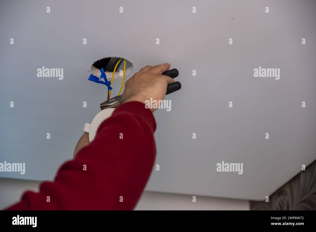 Junger Mann, der den Deckenstrahler repariert, der den Schäler hält, um Drähte zu schneiden und sie mit neuen zu befestigen Stockfoto
