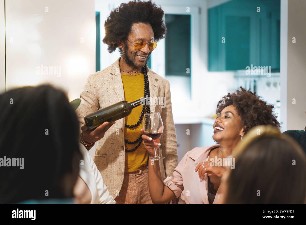 Mann im Retro-Afro-Stil serviert Wein für lachende Freunde bei einem intimen Treffen im Haus - Momente des Genusses und der Zweisamkeit. Stockfoto
