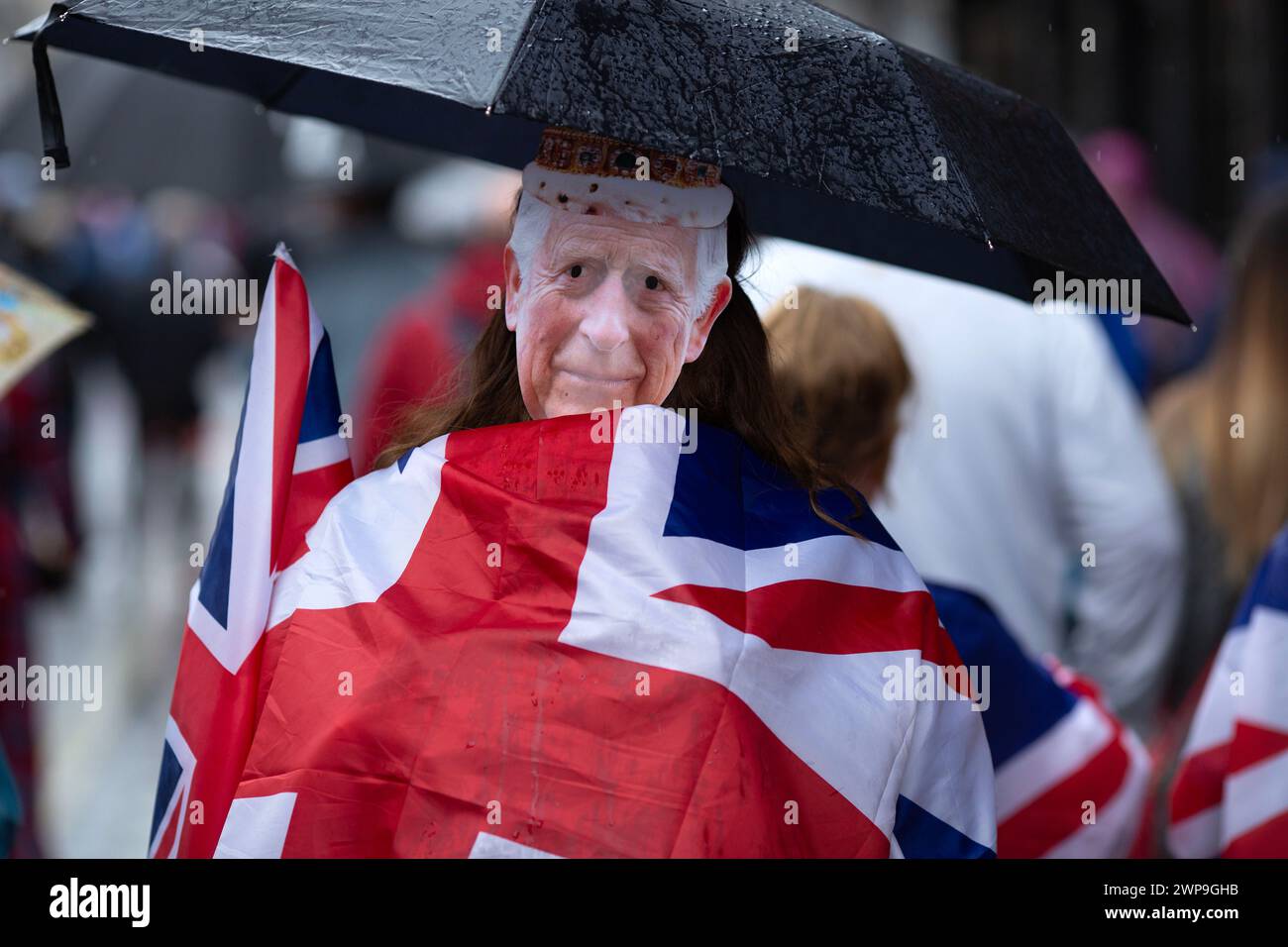 Eine Maske von König Karl III. Ist auf dem Kopf eines Fußgängers zu sehen, der eine Gewerkschaftsflagge trägt, während sich die Menschen versammeln, um die Krönung des Königs in London zu sehen. Stockfoto