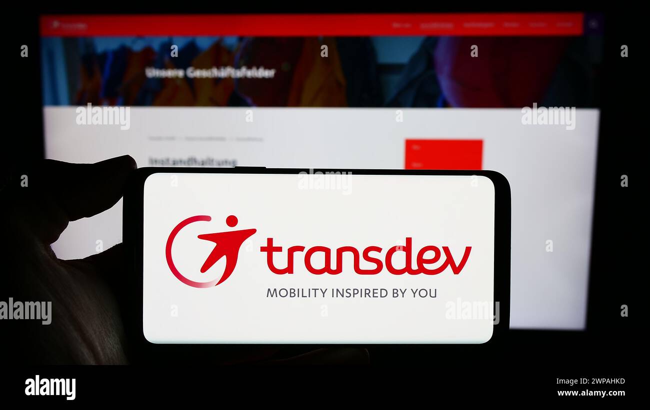 Person, die ein Mobiltelefon mit dem Logo des deutschen Verkehrsunternehmens Transdev GmbH vor der Webseite hält. Schwerpunkt auf dem Display des Telefons. Stockfoto