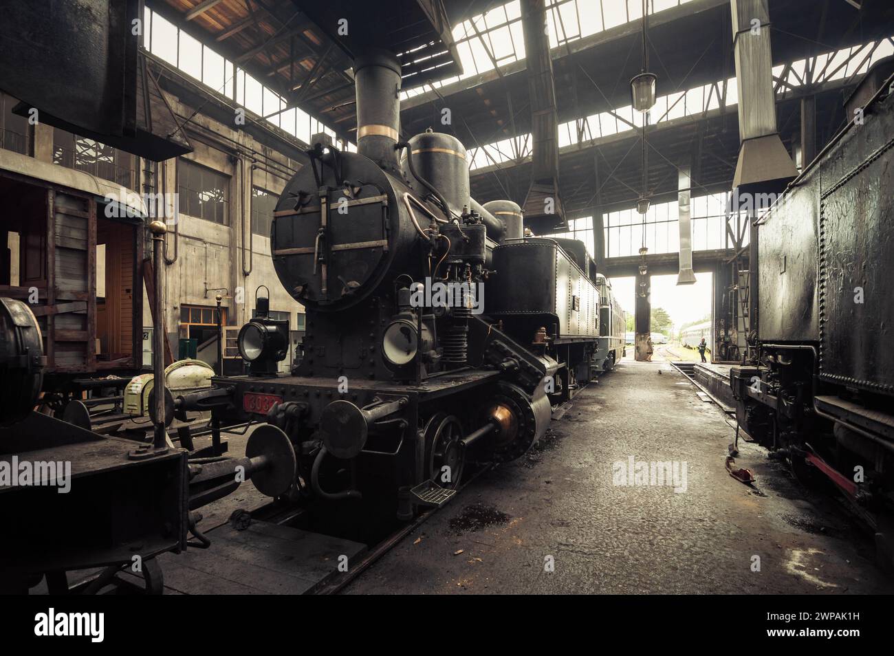 Alte Dampflokomotive kkStB 30 und andere Züge im Bahndepot. Die Luft im Bahnschuppen ist mit nebeligem Rauch gefüllt. Bild in warmen Brauntönen. Stockfoto