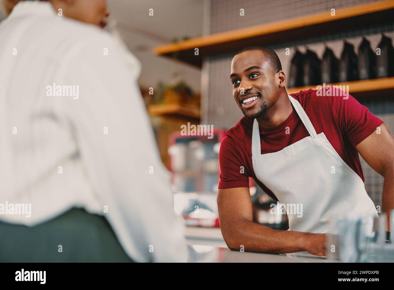 Ein fröhlicher männlicher Barista auf einer Schürze bietet ausgezeichneten Kundenservice in einem modernen Café. Die helle Inneneinrichtung und die angenehme Interaktion zeigen ein warmes, Stockfoto