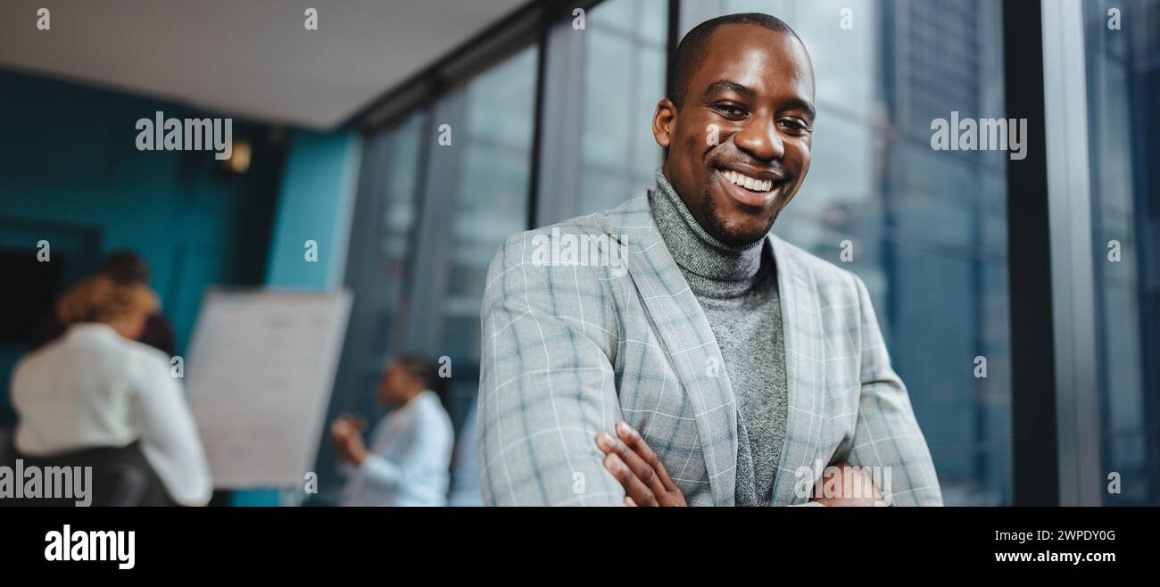 Selbstbewusster afrikanischer Unternehmer steht während eines Geschäftstreffens in einem Konferenzraum voller Menschen. Er trägt einen Anzug, hat Arme überkreuzt und lächelt ihn an Stockfoto