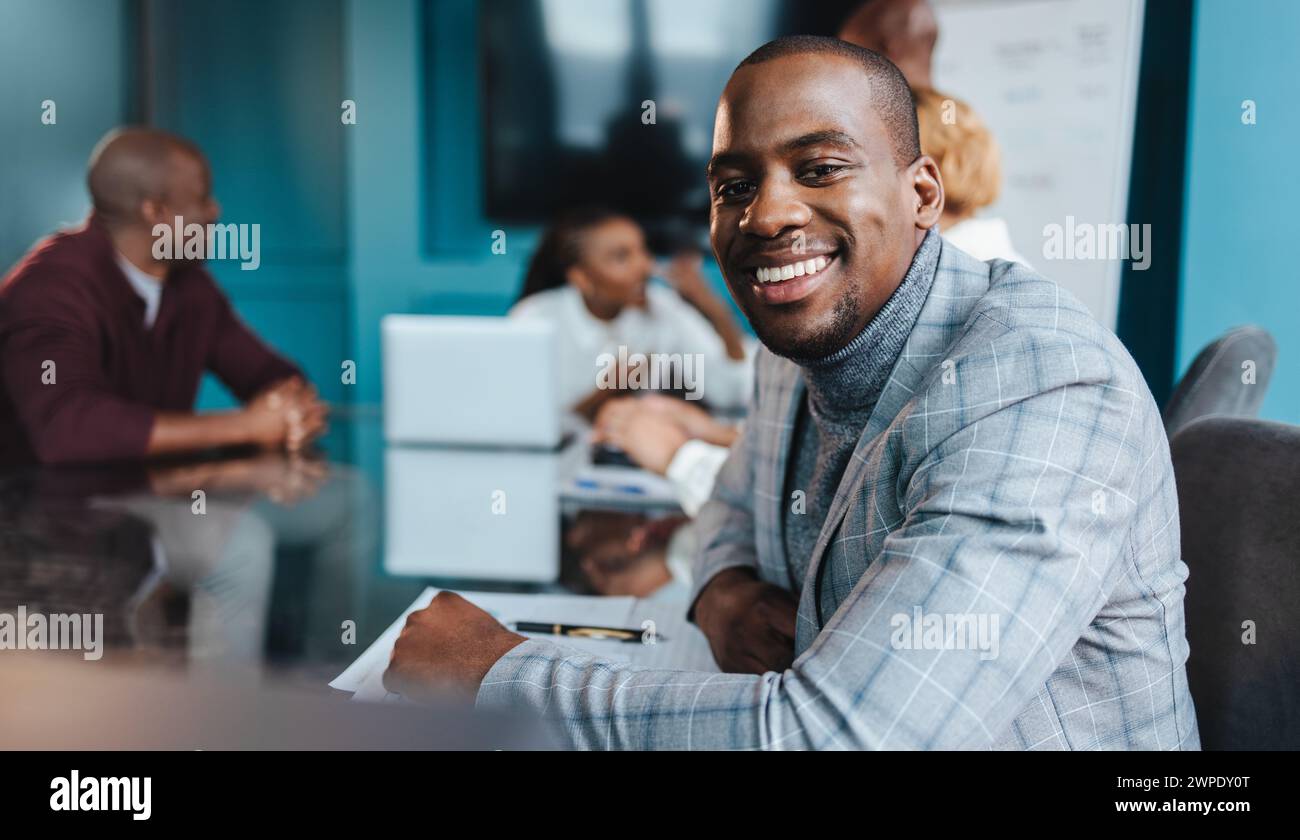 Ein professioneller Geschäftsmann in einem stilvollen Anzug zeigt Selbstvertrauen mit einem warmen Lächeln während eines Teammeetings in einem modernen Büroset Stockfoto