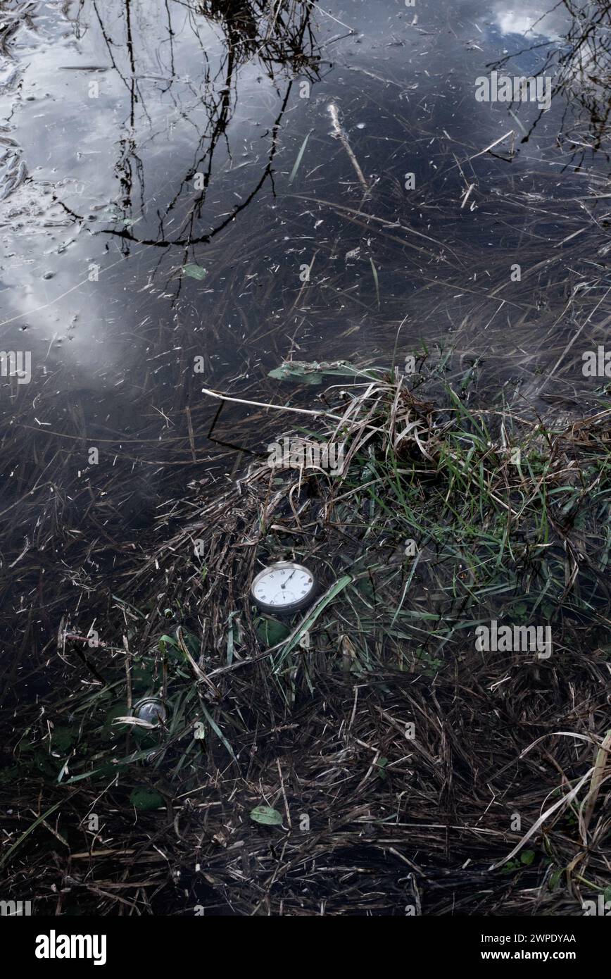 Eine alte silberne Taschenuhr, die im flachen Wasser am Rand eines Teichs liegt, Wolken spiegeln sich im Wasser. Stockfoto