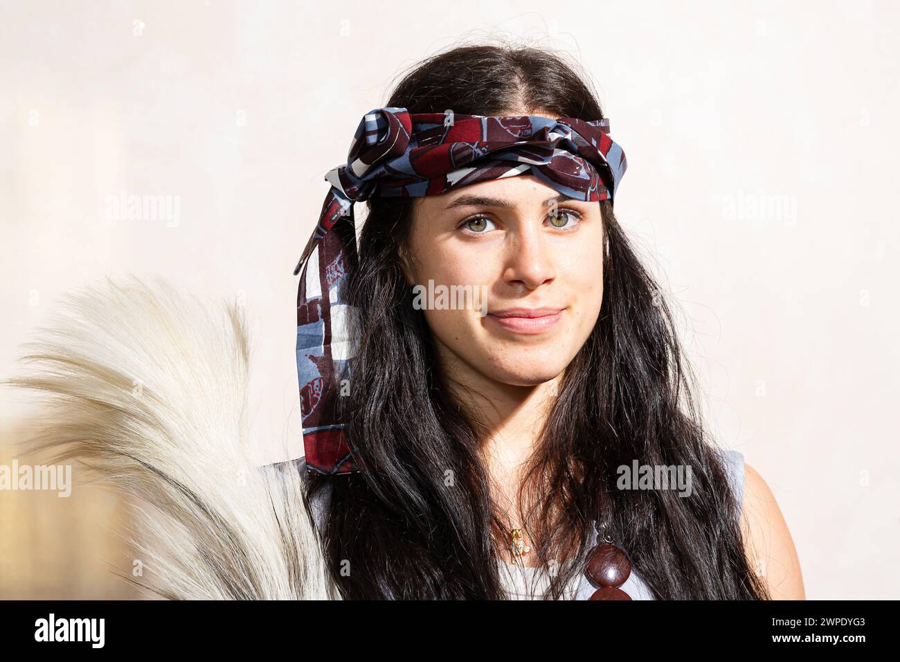 Nahaufnahme einer fröhlichen jungen Frau mit gemustertem Kopftuch und weißem Tanktop, die einen traditionellen Rosshaar-Fan hält. Die warme Innenbeleuchtung hig Stockfoto