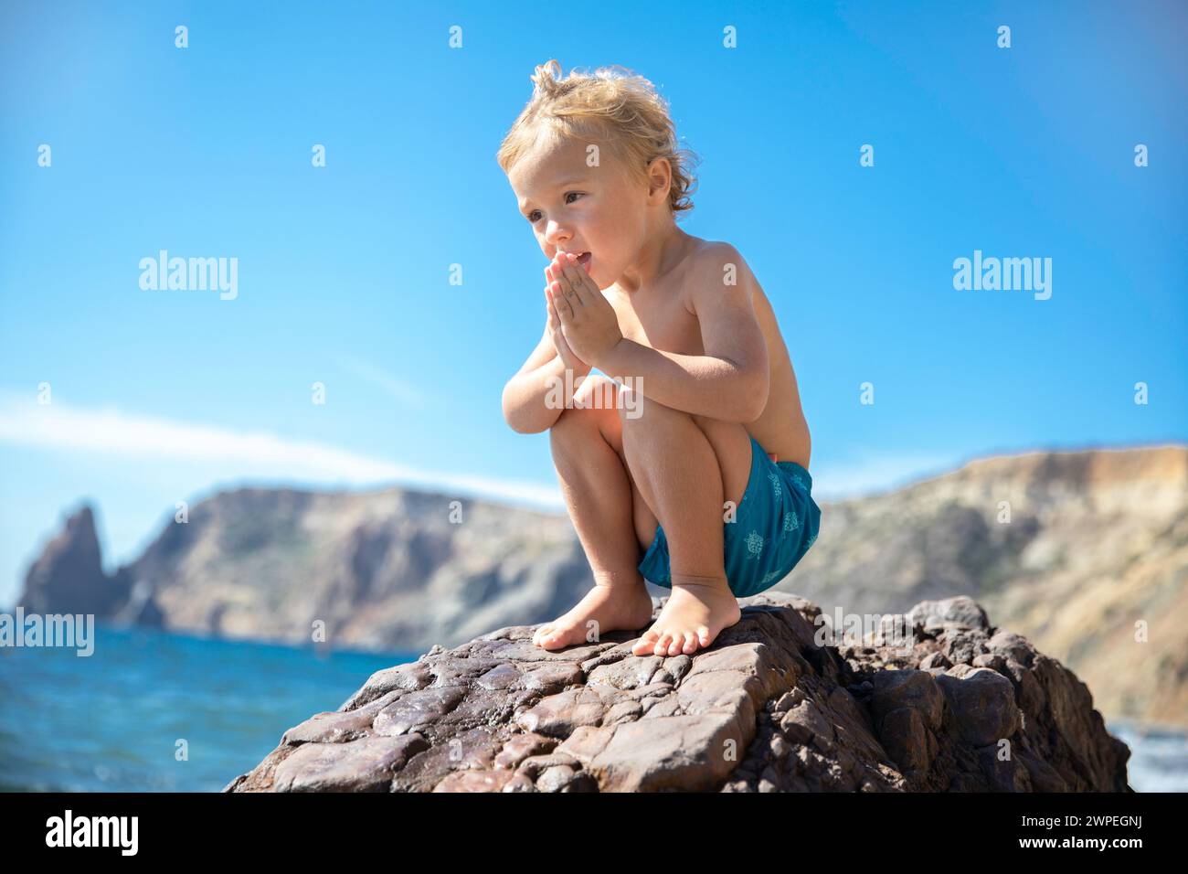 Ein kleiner blonder Junge in Badehosen spielt an einem warmen sonnigen Tag auf einem Stein am Meer. Stockfoto