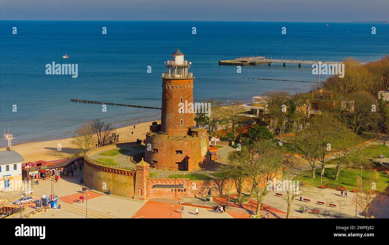 Februar in Kołobrzeg, Polen: Eine ruhige Szene mit einem Leuchtturm aus rotem Backstein, Menschen, die einen sonnigen Tag am Sandstrand genießen, und einem ruhigen Meer mit Stockfoto