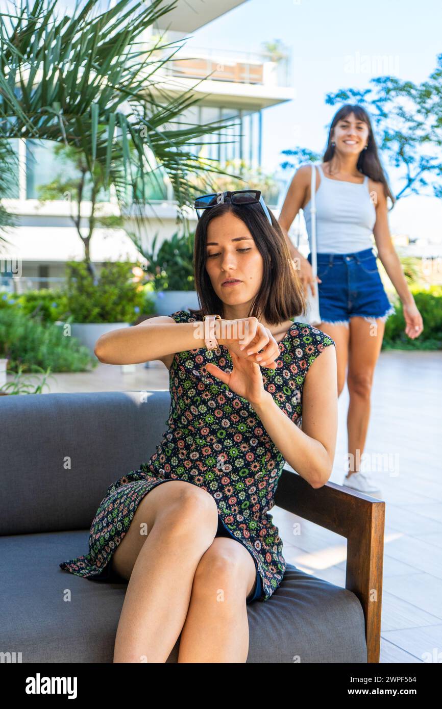 Eine Frau sitzt auf einer Couch und schaut auf ihre Uhr und wartet auf einen Freund, der hinter ihr steht Stockfoto