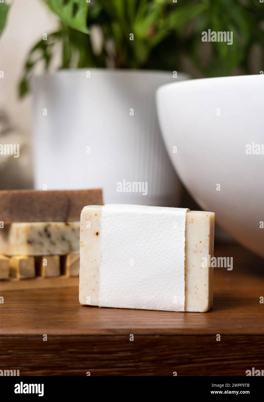Beige handgemachte Seifenstange mit leerem Etikett auf der hölzernen Arbeitsplatte im Badezimmer in der Nähe von Topfmonstera Pflanze und Becken in Nahaufnahme, Verpackungsmodell, Kopierraum. Na Stockfoto