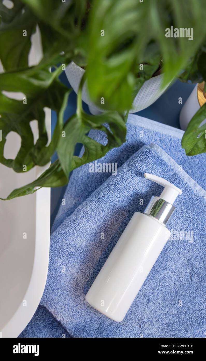 Weiße Kosmetikflasche mit einer Pumpe auf gefalteten blauen Handtüchern in der Nähe des Waschbeckens und der grünen Monstera, Blick von oben, Mockup. Lifestile Szene mit Hautpflege-Schönheitsprodukt in Stockfoto
