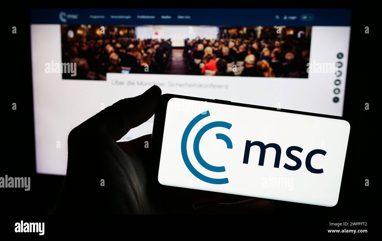 Person mit Smartphone mit Logo der jährlichen Münchner Sicherheitskonferenz (MSC) vor der Website. Schwerpunkt auf dem Display des Telefons. Stockfoto