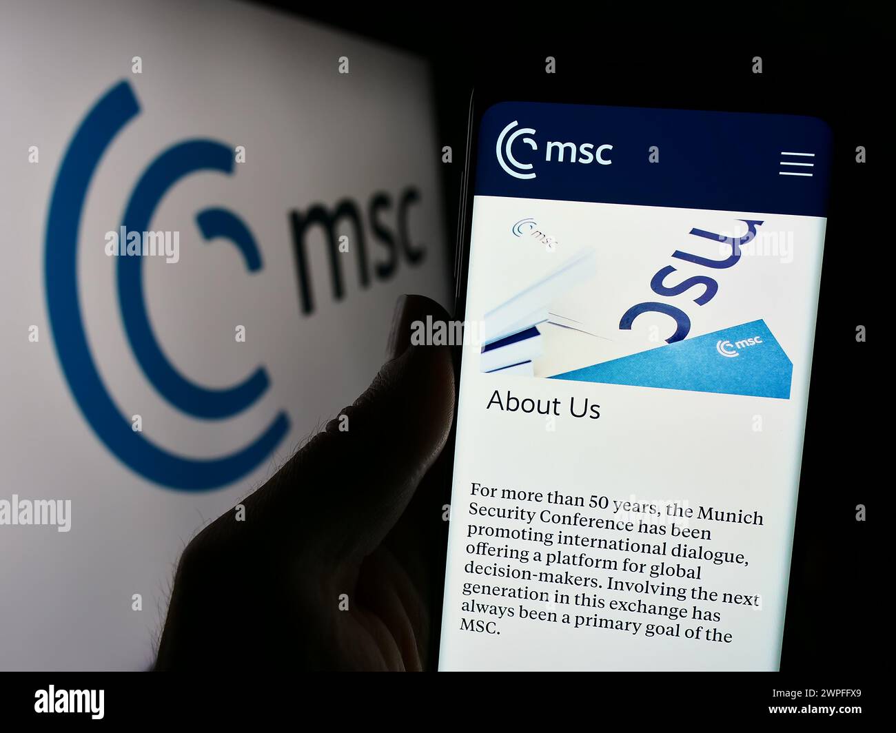 Person mit Mobiltelefon mit Webseite der jährlichen Münchner Sicherheitskonferenz (MSC) vor Logo. Konzentrieren Sie sich auf die Mitte des Telefondisplays. Stockfoto