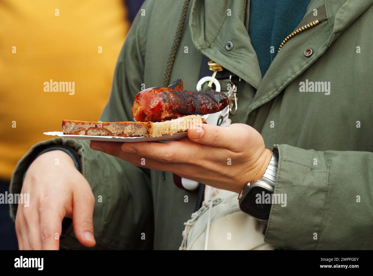 Mitte eines Kunden, Besucher des Bauernmarktes mit Papiertablett mit Snack, Brotscheibe mit gegrillter Wurst in der Hand. Nahaufnahme, lange Zeit Stockfoto