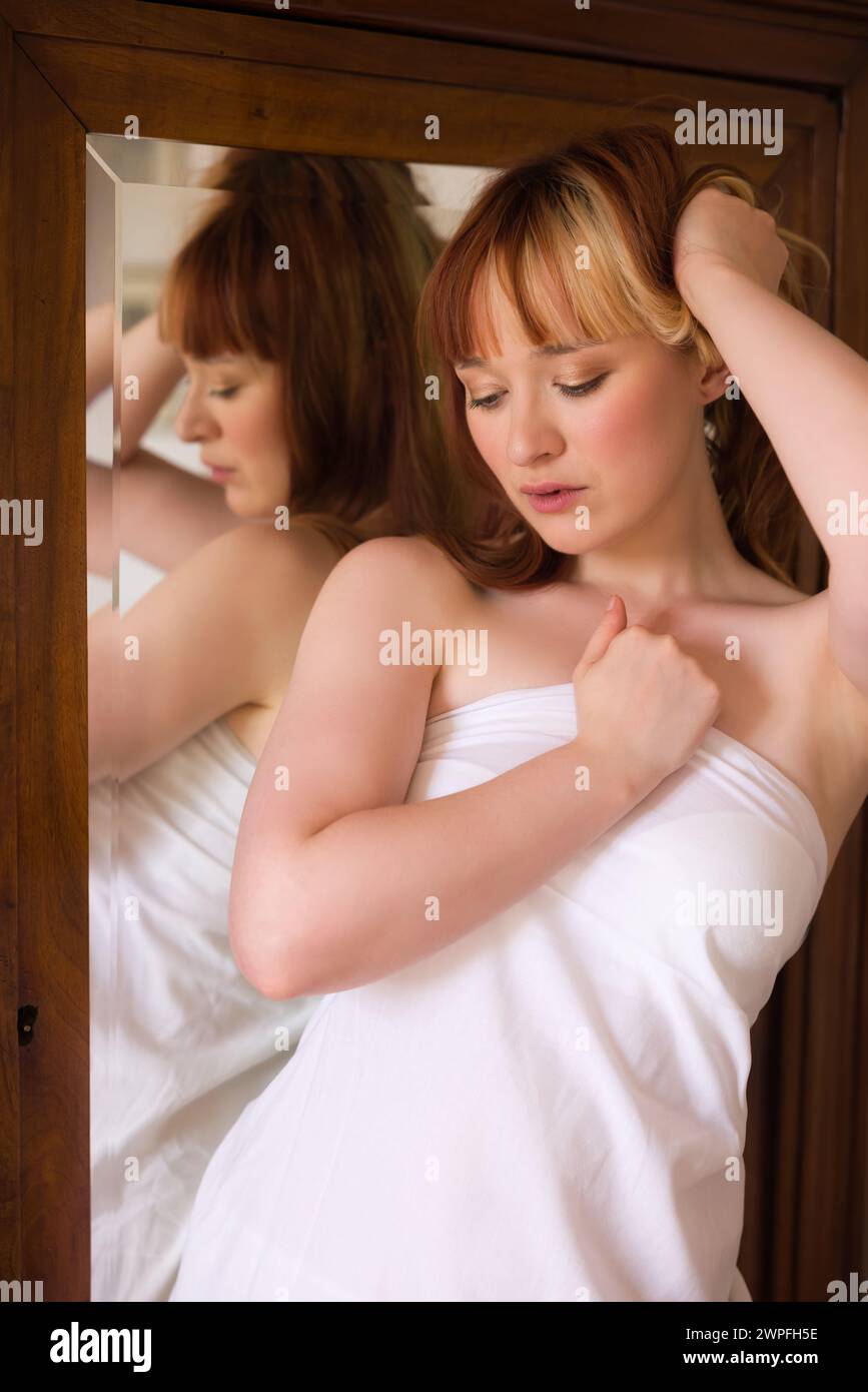 Attraktive junge Frau, die mit einem weißen Bettlaken vor einem Spiegel im Kleiderschrank posiert Stockfoto
