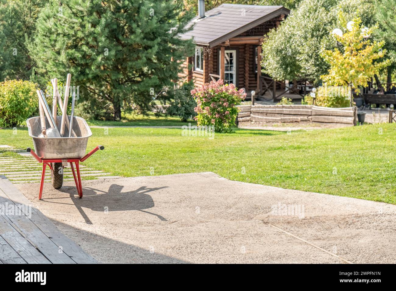 Metallwagen mit Gartengeräten im sonnigen Garten auf dem Hinterhof des Landhauses. Wagen mit Handinstrumenten des Gärtners auf dem Rasen des Landhauses. Landleben Stockfoto