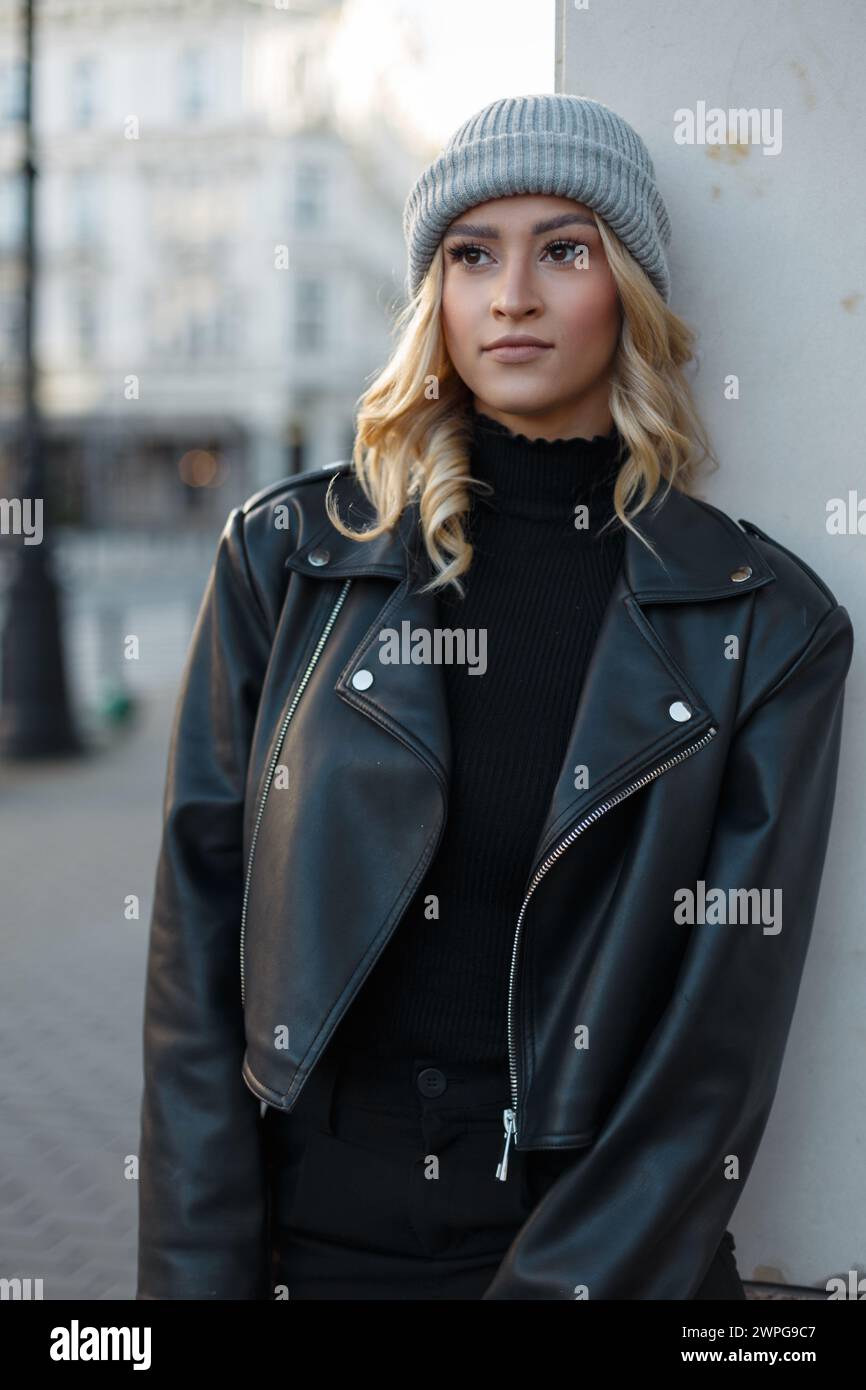 Urbanes modisches, hübsches, schickes Mädchen mit blondem Haar und Hut in schwarzer Casual Rock Kleidung mit Lederjacke und Pullover in der Stadt. Niedliche Frau Stockfoto