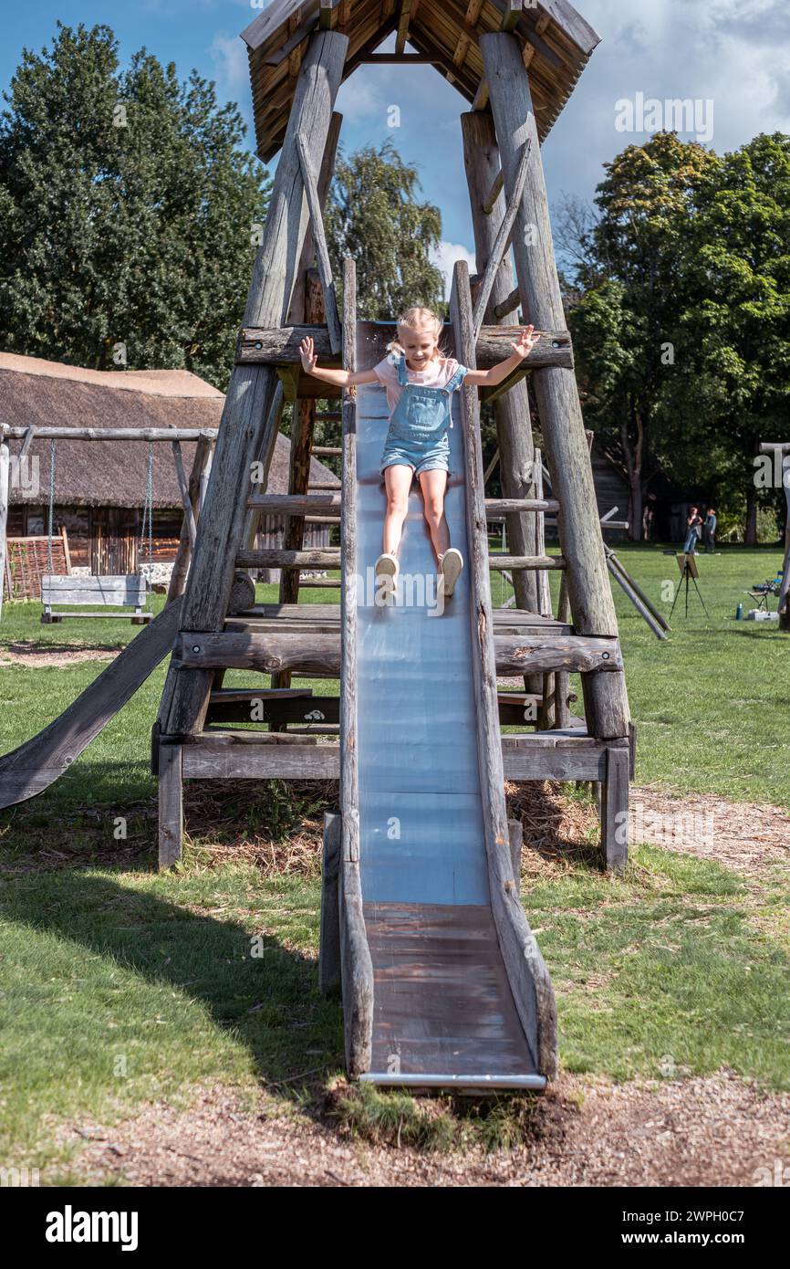 Ein Mädchen im Jeansanzug rutscht fröhlich auf einem Spielplatz auf einer Holzrutsche hinunter und verkörpert Freiheit und Verspieltheit vor einem bewölkten Himmel, Krankenkasse Stockfoto